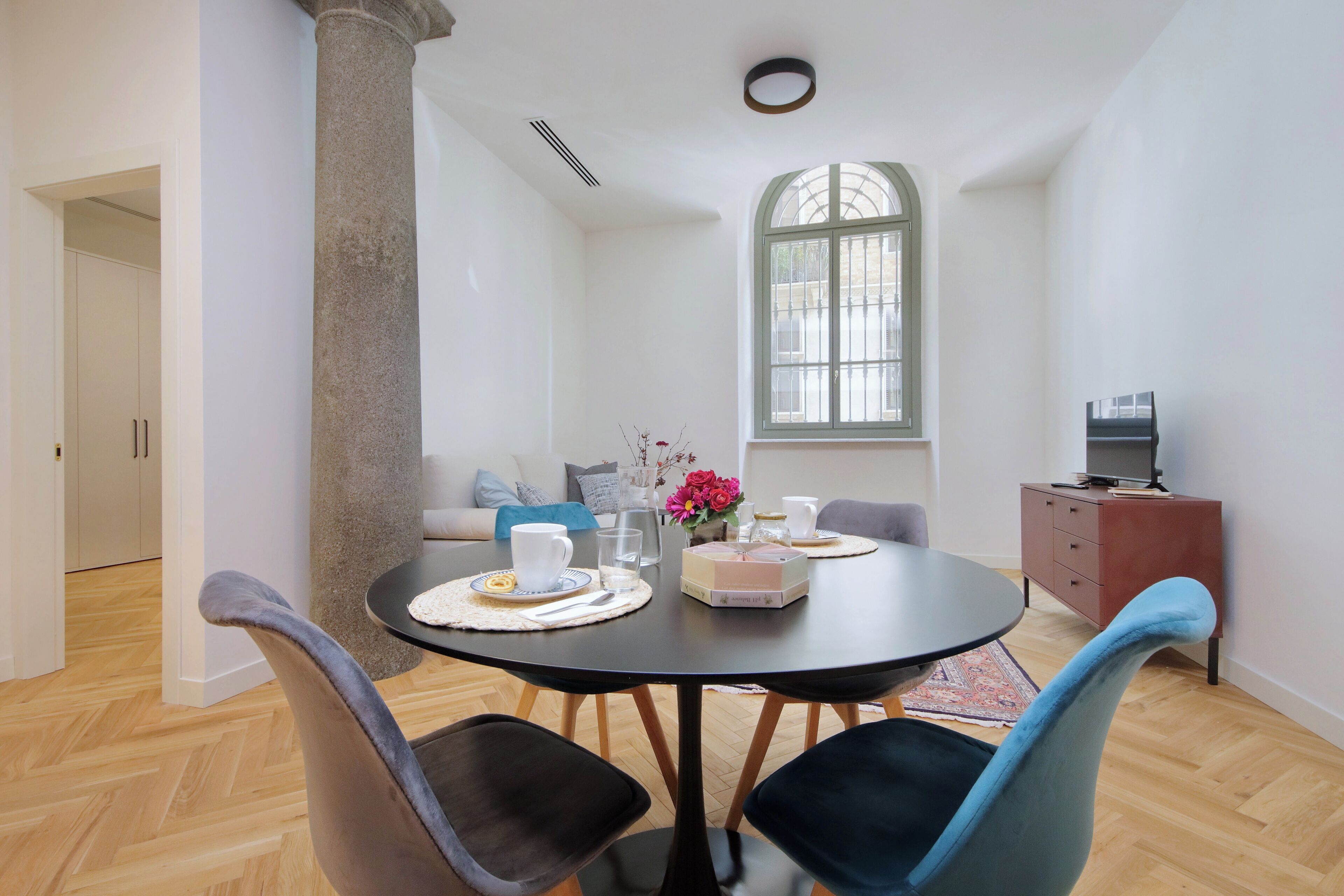 Un comedor con una mesa redonda, sillas, un jarrón con flores y un televisor sobre un mueble.