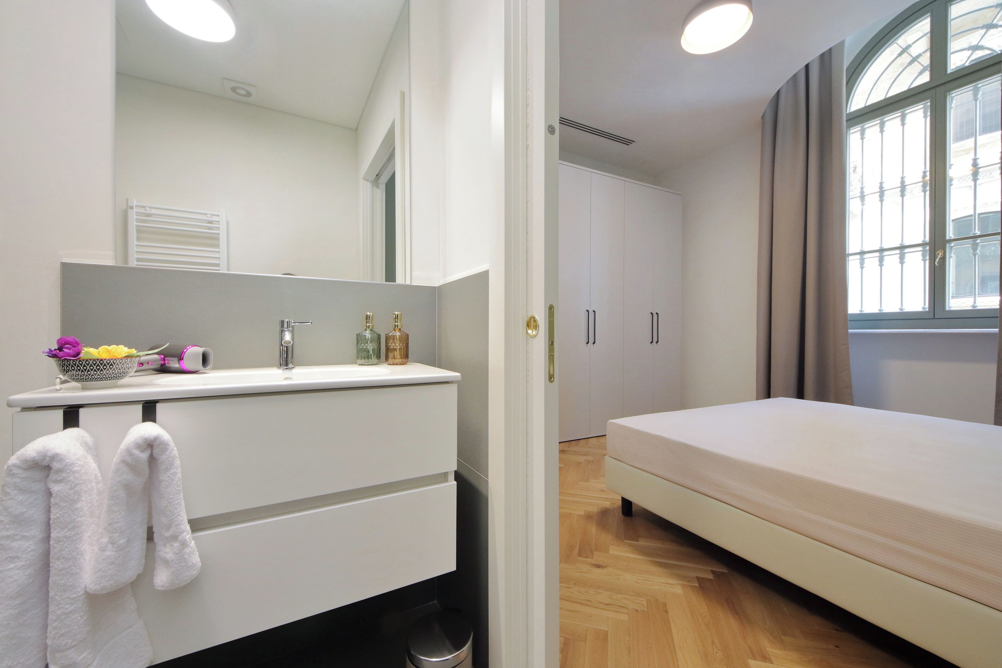A bedroom with a bed, a bedside table with a mirror, a window with curtains, and a bathroom with a sink and towel rack.
