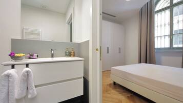 A bedroom with a bed, a bedside table with a mirror, a window with curtains, and a bathroom with a sink and towel rack.