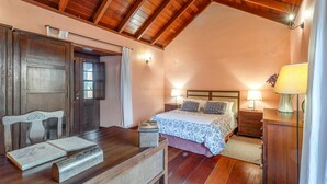 A bedroom with a wooden ceiling, a bed with patterned bedding, a nightstand, a dresser, and a chair with an open book. - Country House 'Gran Drago' with Sea View, Wi-Fi and Air Conditioning (Villa de Valverde)