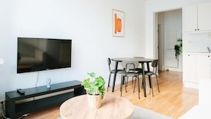 Uma sala moderna com televisão de tela plana, uma mesa de jantar com cadeiras, uma pequena planta em vaso e uma área de cozinha com armários.