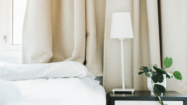 Cama bem arrumada com lençóis brancos, abajur e uma pequena planta em vaso num criado-mudo.