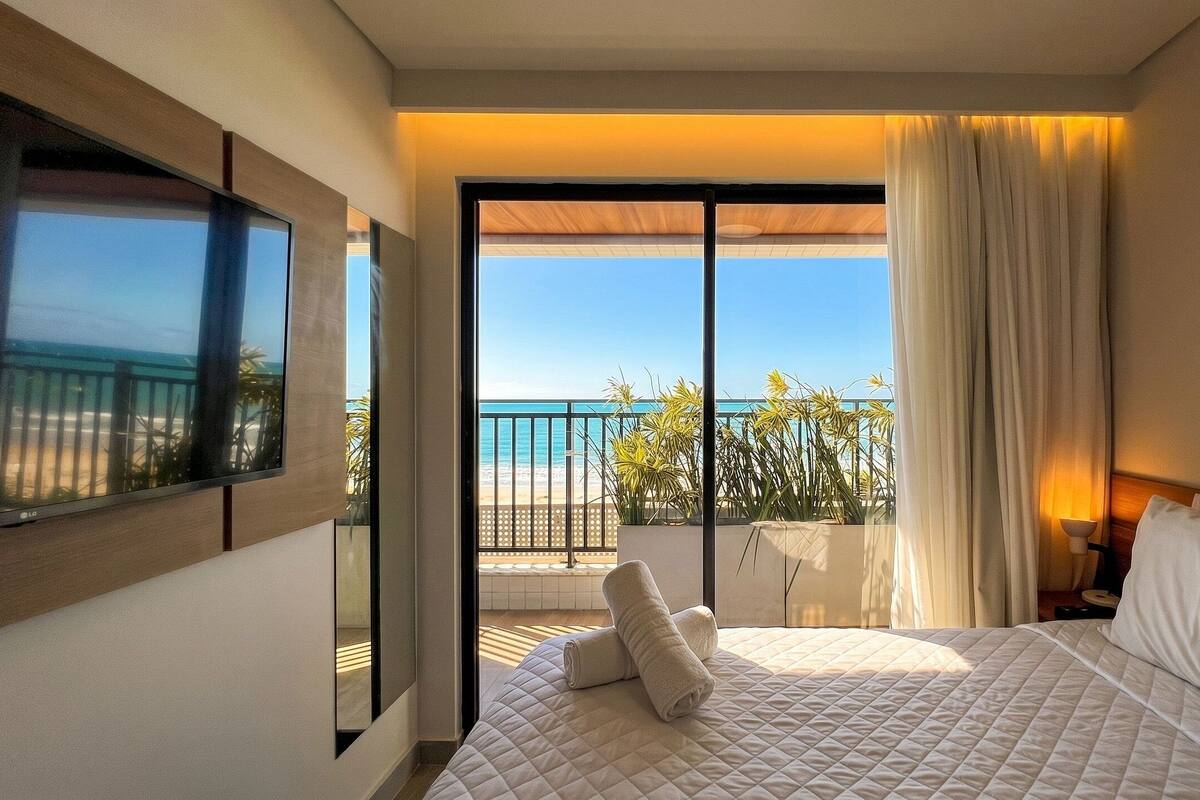 Une chambre d’hôtel avec un lit, une vue sur la plage et un balcon agrémenté de plantes.