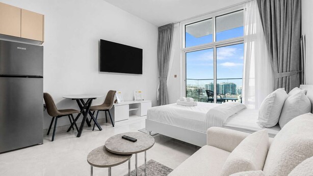 A modern hotel room with a bed, a seating area with chairs, a dining table, a TV, and a large window with a city view. - StayShort - Premium New Arjan Studio - sleeps 3 (Arjan)