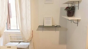 A room with a window, a radiator, a bed, and two wall-mounted shelves with a plant and a framed document. - Colosseum: Exposed Brick Double & Private Bath (Roma)