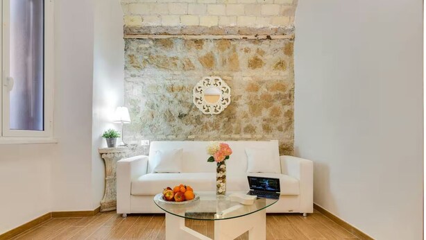 A living room with a white sofa, a glass coffee table with fruit, a laptop, and a decorative mirror on a brick wall. - Colosseum: Exposed Brick Double & Private Bath (Roma)
