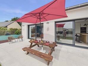 An outdoor dining area with a wooden table and benches, a red umbrella, and a view of the house's interior through glass doors. - New house for 6 with garden and terrace (Plonéour-Lanvern)