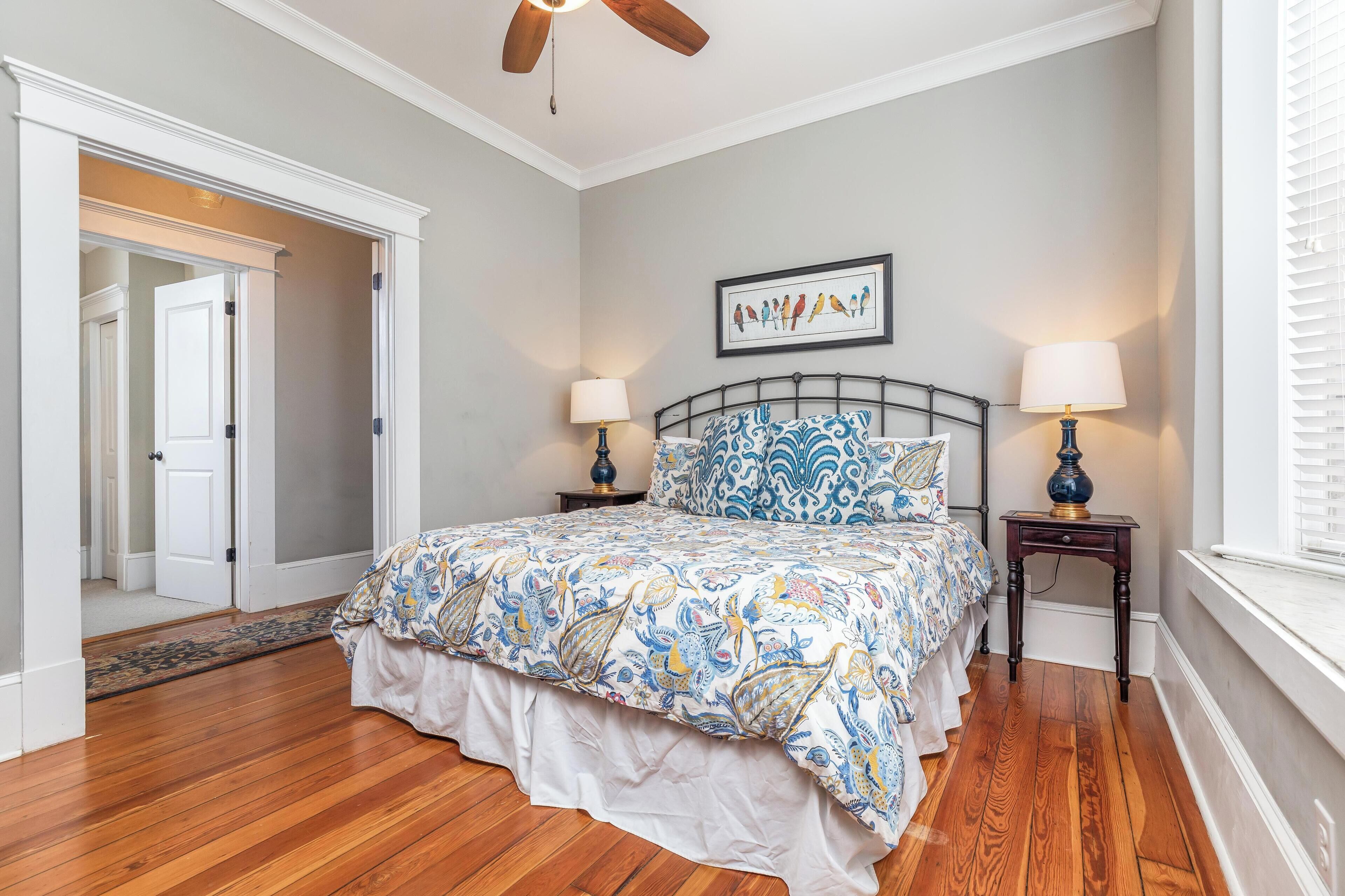 Une chambre à coucher comprenant un lit, deux tables de chevet, un ventilateur de plafond, un tableau encadré au mur et une fenêtre avec des stores.