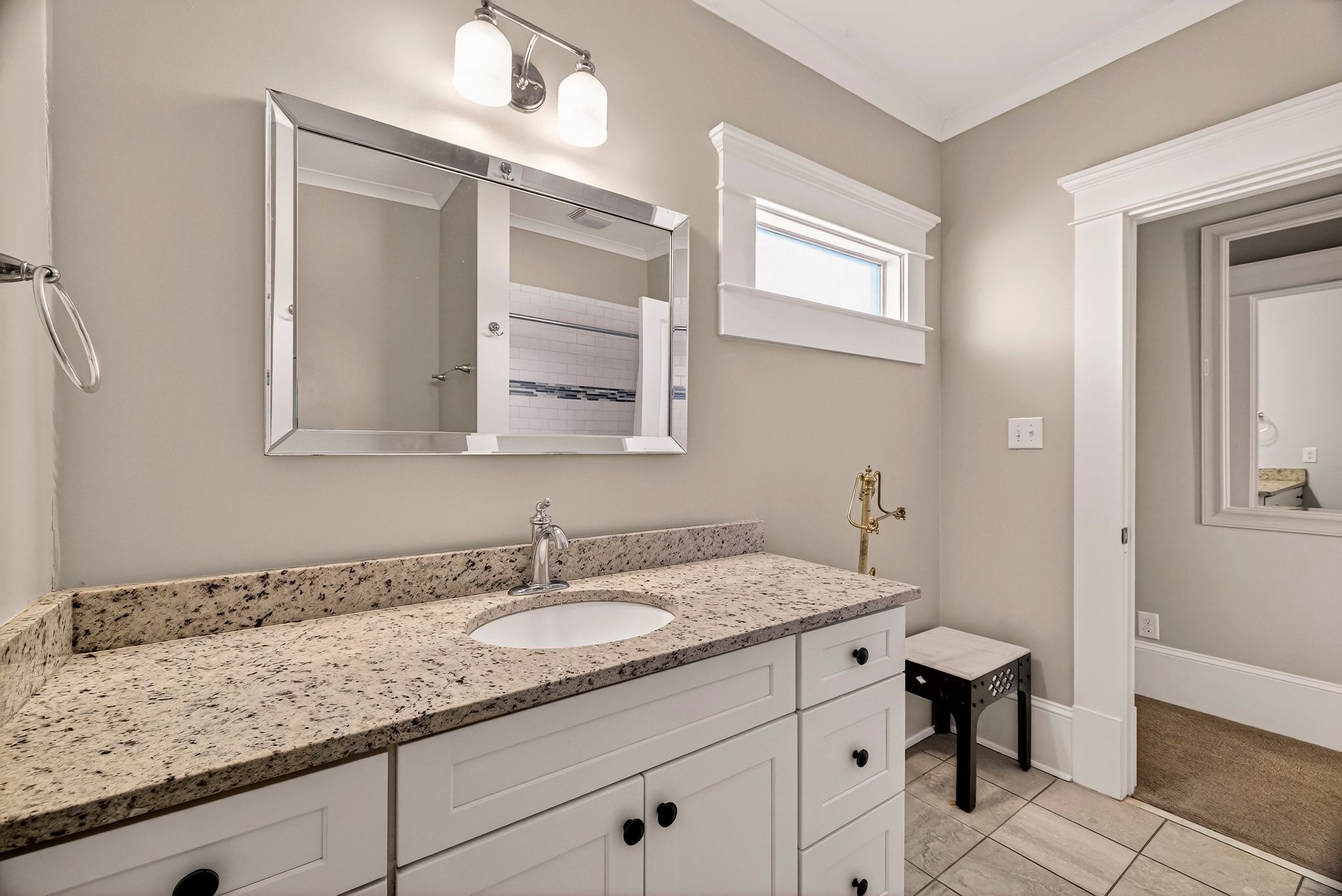 Une salle de bain avec un lavabo surmonté d’un plan de travail en granit, des meubles blancs, un grand miroir et un petit tabouret.
