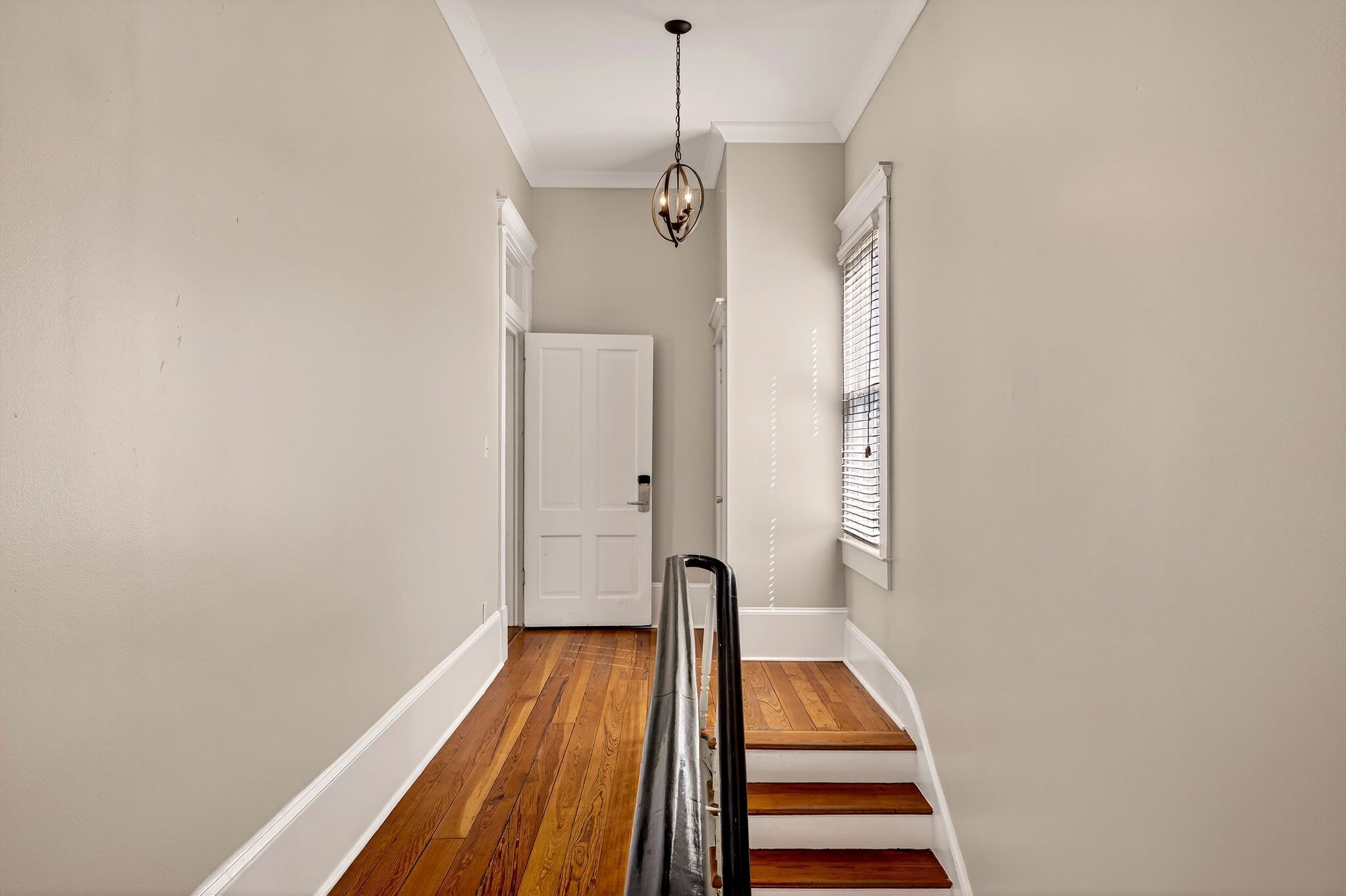 Un couloir doté d’un plancher en bois, de murs blancs, d’une porte blanche et d’un escalier avec une rampe noire.