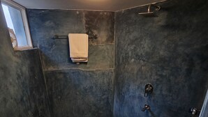 Un baño pequeño y de color oscuro con una toalla colgada en la pared, un ventanal visible y un panel de control de la ducha.