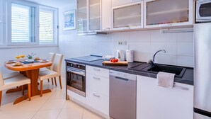 A modern kitchen with white cabinets, a built-in oven, a dishwasher, and a dining area with a table and chairs. - Apartment Promenada (Dubrovnik)