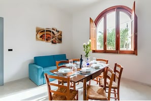 A dining area with a table set for six, a blue sofa, wooden chairs, a large window with an open door, and a wall-mounted abstract art piece. - Villa Anny with Hot Tub Just Steps from the Sea (Avola)