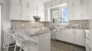 Una cucina moderna con mobili bianchi, zoccolo in piastrelle, un'ampia finestra e un angolo bar con sgabelli.