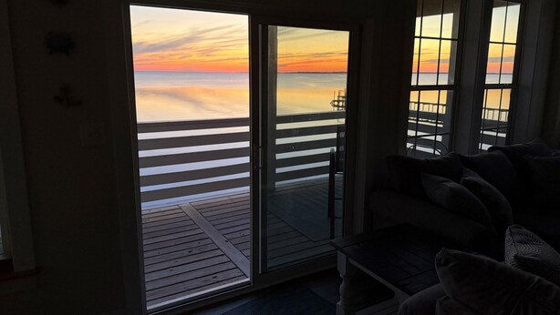 A room with a view of a sunset over a pier, a sliding glass door, and a dark-colored sofa. - Waterfront Seclusion Stunning Sunsets and Golf! (Jarvisburg)