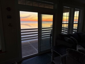 Een kamer met uitzicht op een ondergaande zon boven een pier, een glazen schuifdeur en een donkergrijze bank.