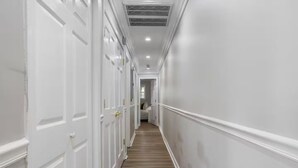 Um corredor estreito com paredes brancas e soalho de madeira, que leva a um quarto com cama e janela.