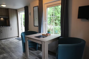 A modern room with a wooden table, two chairs, a TV mounted on the wall, and a window with a view of greenery.