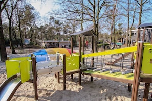 A playground with various play structures, including a slide and climbing frames, set in a sandy area with trees in the background.