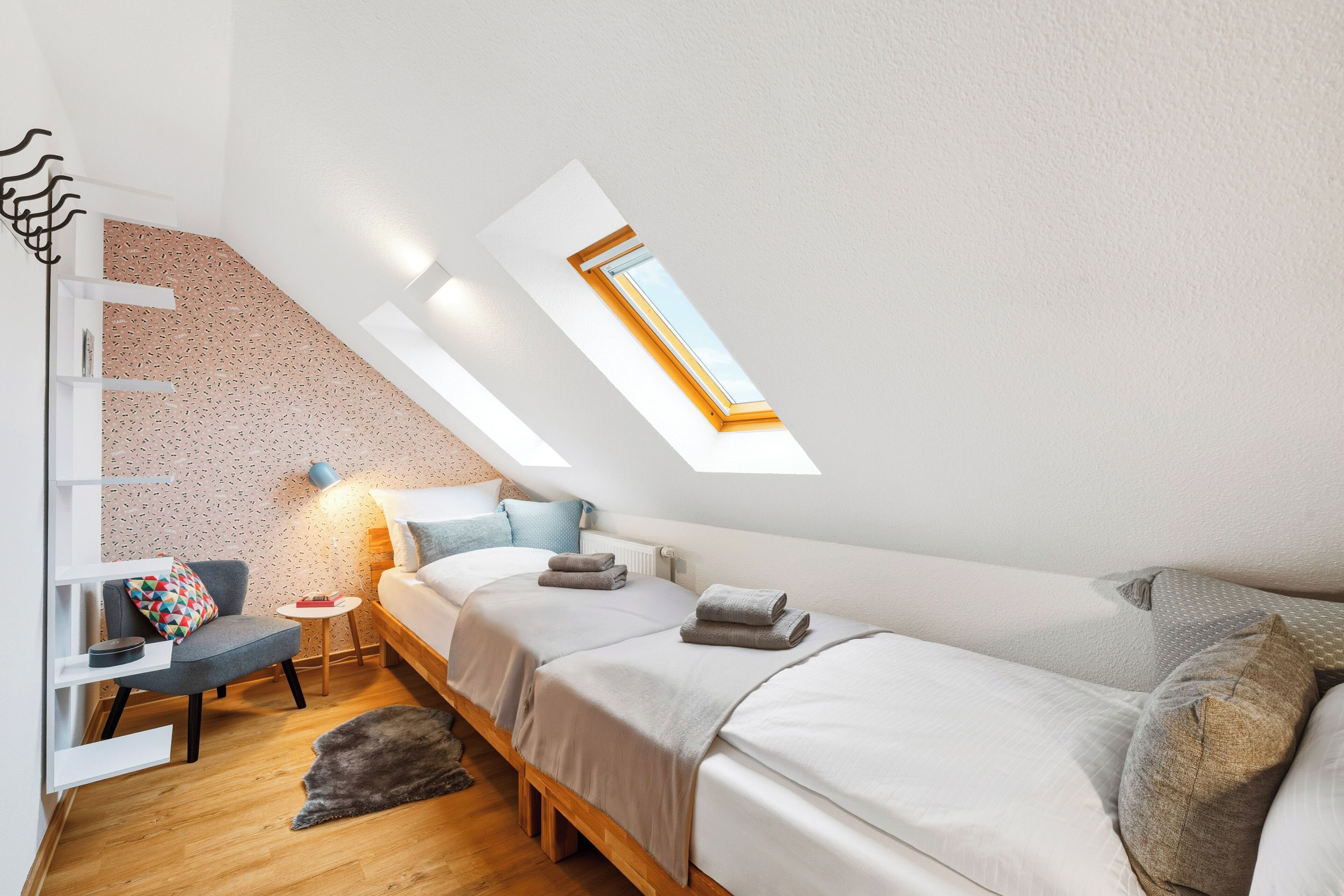 Ein Schlafzimmer mit schrägem Dach, Oberlichtern, einem Bett, einem Sessel, einem kleinen Tisch und einer Wanddekoration.