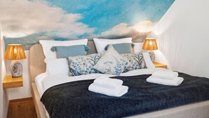 Ein ordentlich bezogenes Bett mit dunkler Decke, weißen Kissen und einer blumigen Kissenanordnung in Blau und Weiß. Auf den hölzernen Nachttischen stehen jeweils eine Nachttischlampe mit beigefarbenem Lampenschirm.