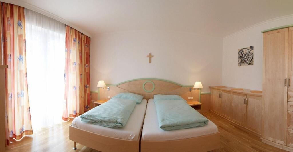 A double bed with two pillows, a wooden headboard, two bedside lamps, a wooden wardrobe, and a cross on the wall.