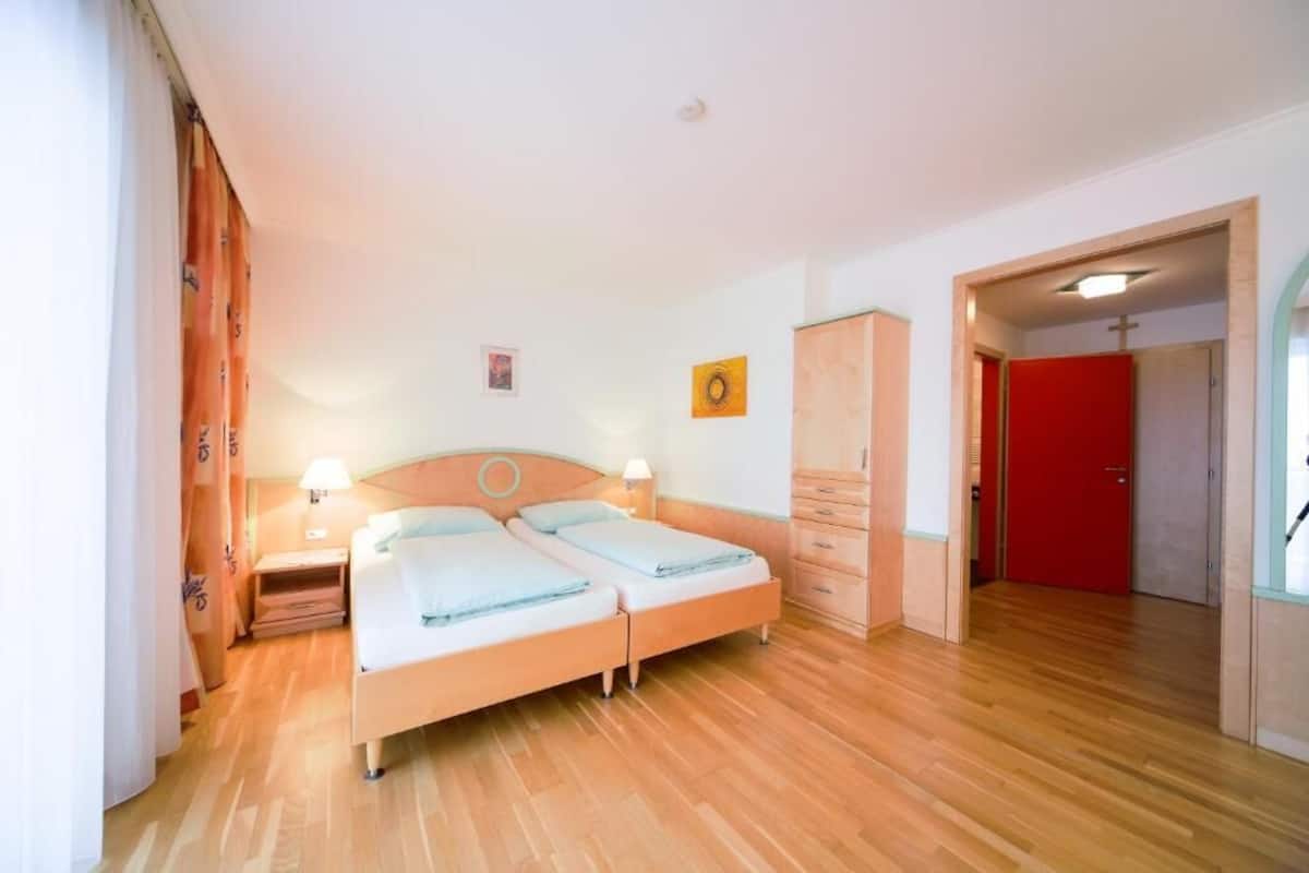 Uma cama de casal com duas mesas de cabeceira, um guarda-roupa e uma porta vermelha num quarto de hotel.