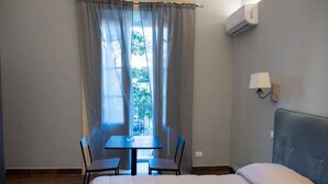 Een kamer met een bed, een tafel met twee stoelen, een raam met gordijnen, een airconditioningapparaat en een wandlamp.