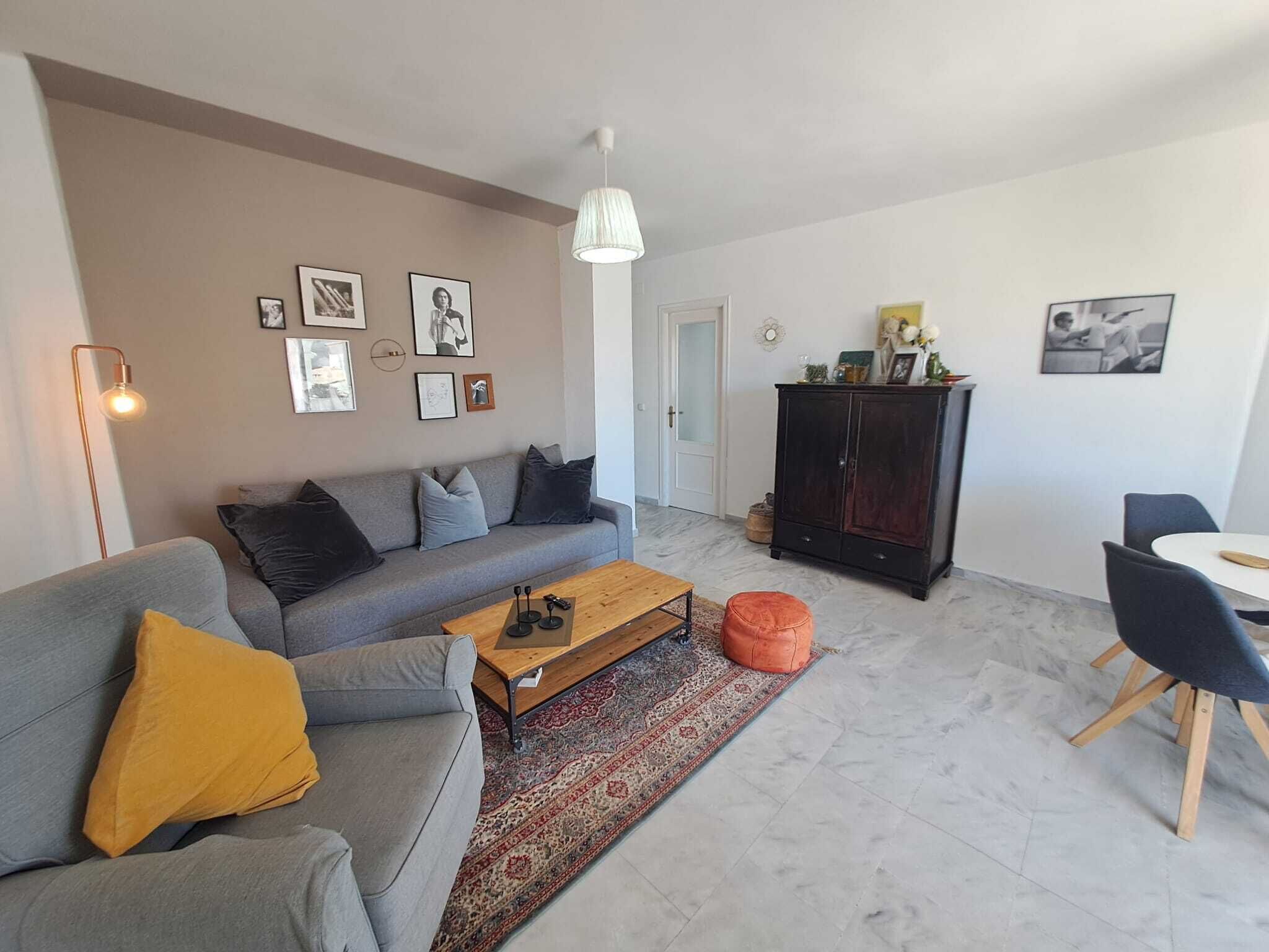 Una sala moderna con un sofá gris, una mesa de centro de madera, un mueble oscuro y un comedor con mesa y sillas blancas.