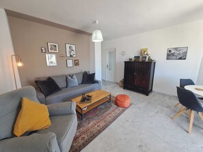 Ein modernes Wohnzimmer mit einem grauen Sofa, einem Holztisch, einer dunklen Kommode und einem Essbereich mit weißem Tisch und Stühlen.