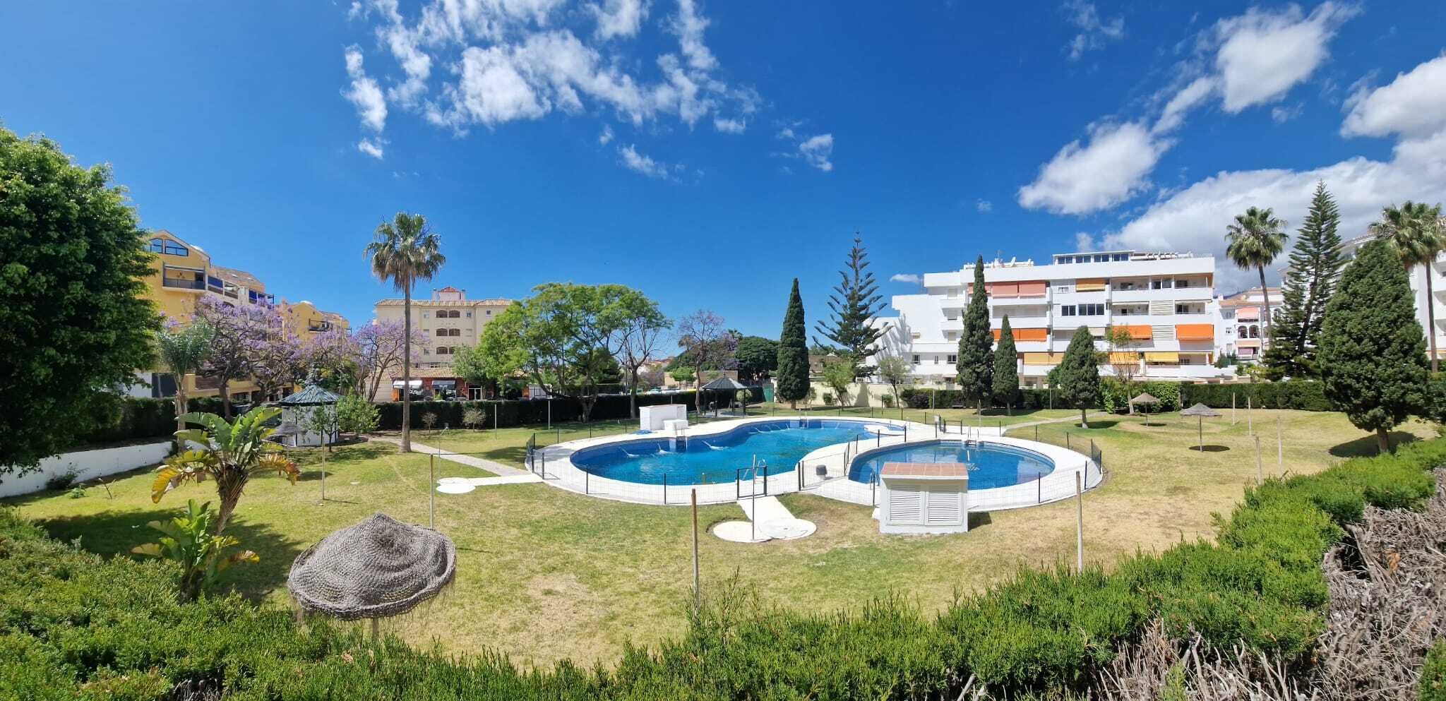 Una piscina rodeada de césped, árboles y un edificio de varios pisos al fondo.