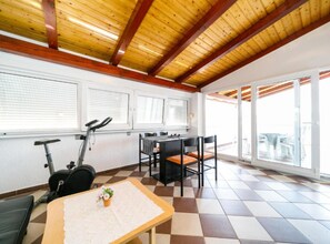 A room with a checkered floor, a table with a lace tablecloth, a small potted plant, an exercise bike, and a dining table with chairs.