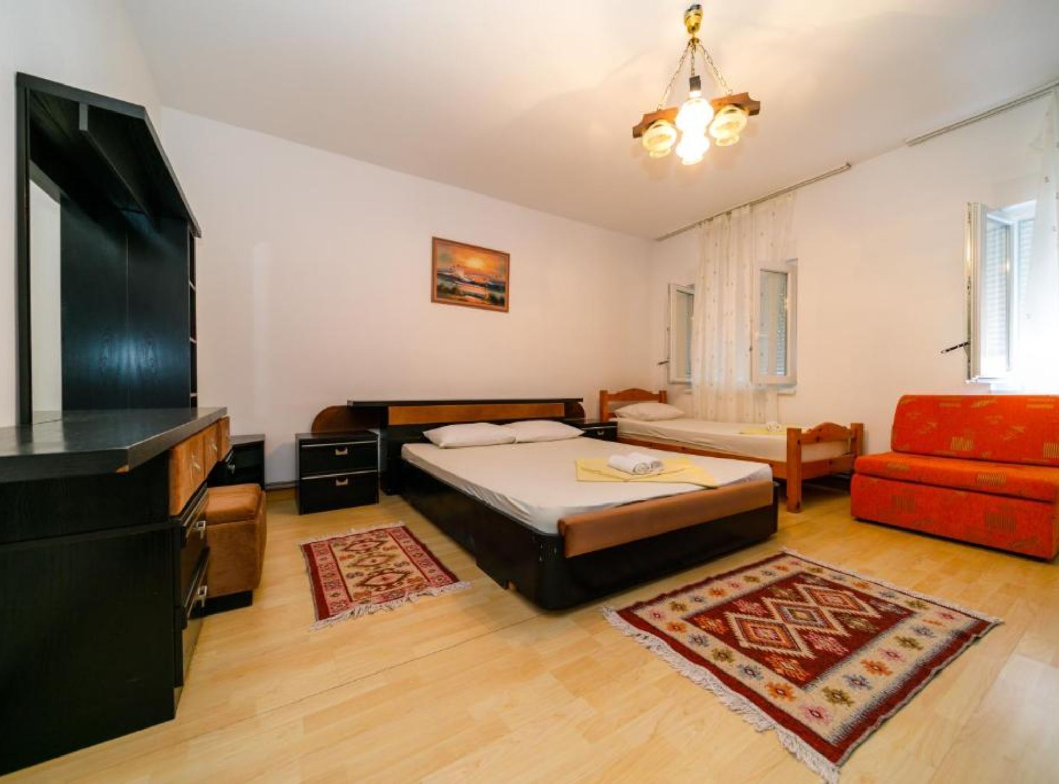 A bedroom with a bed, two bedside tables, a dresser, a sofa, a window with curtains, a painting on the wall, and a chandelier.