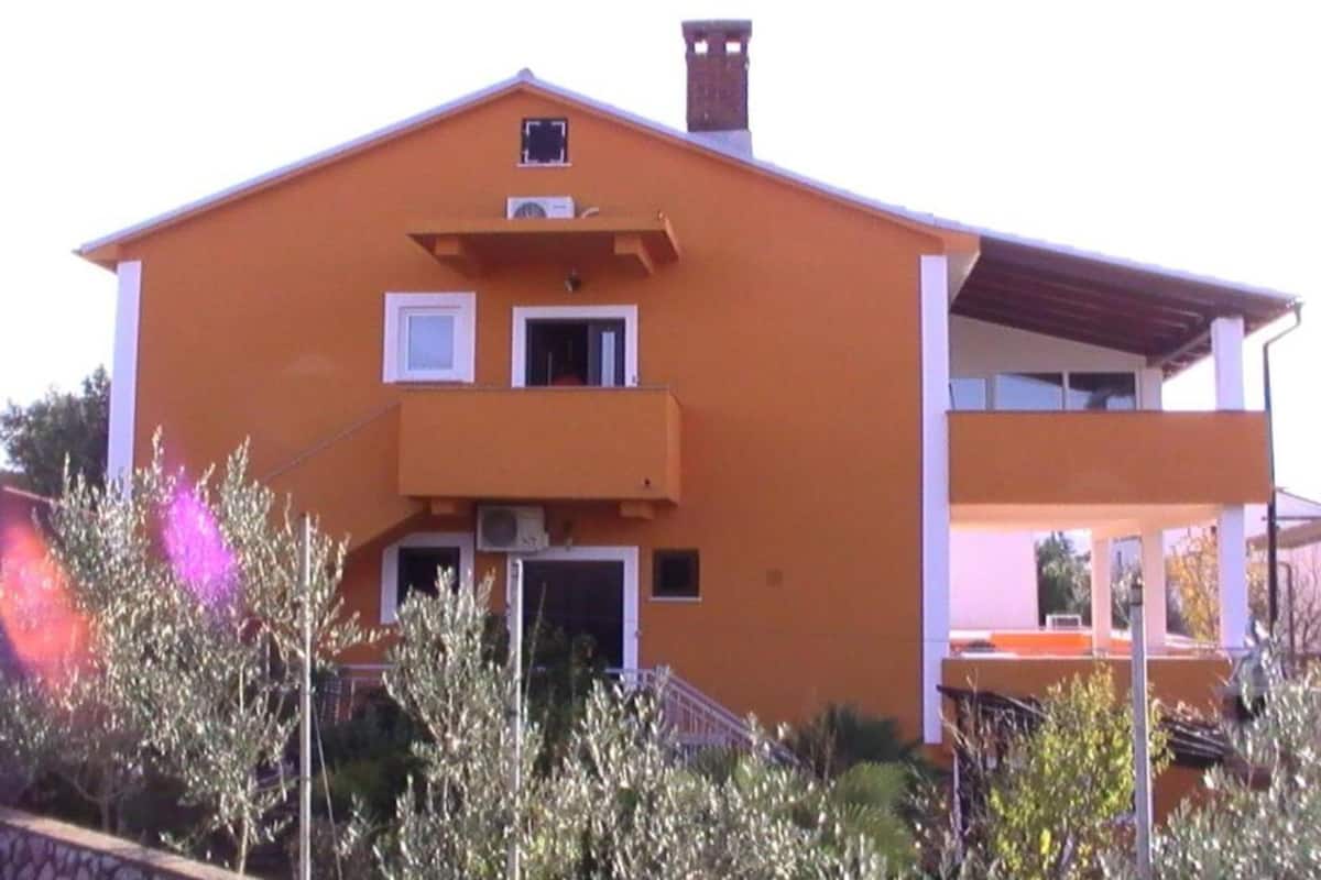 A two-story building with a flat roof, a balcony on the second floor, and a chimney on the roof.