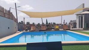 A pool area with a laptop on a wooden table, surrounded by lounge chairs and a shaded canopy. - Room-serene Apartment Escape in Achada F (Santa Cruz)