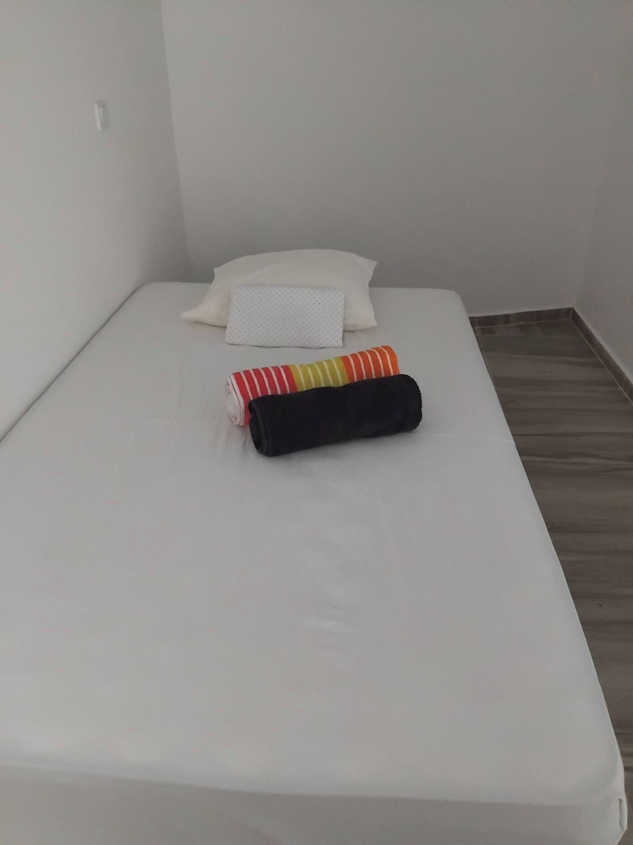 A small, single-bed room with a white mattress, a white pillow, and two rolled-up blankets.