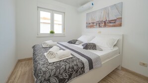 Ein ordentlich bezogenes Bett mit gemusterten Bettwäsche und gerollten Handtüchern, ein Fenster mit Blick auf Gebäude und ein Gemälde mit Segelbooten an der Wand.