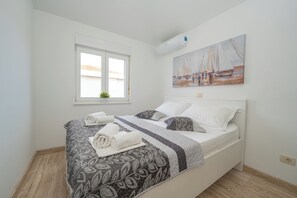 A neatly made bed with patterned bedding and rolled towels, a window with a view of buildings, and a painting of sailboats on the wall. - Two-bedroom apartment Pulse with poolside patio and sea view (Novalja)