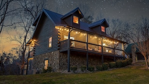 A log cabin with a stone foundation, a balcony with string lights, and a starry night sky.