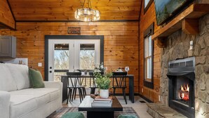 A cozy cabin interior with a stone fireplace, wooden walls, a dining area, and a comfortable seating space.