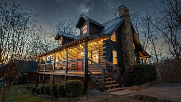 A log cabin with a balcony, stone chimney, and string lights.