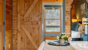 A cozy bedroom with a large wooden door, a window with a view, a bed with a knitted blanket, and a bedside table with a lamp and flowers.