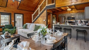 A wooden cabin interior with a dining area, kitchen, and a staircase leading to an upper level.