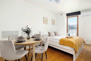 A modern, well-lit room with a dining table, chairs, a bed with a yellow blanket, and a large window offering a city view. - Modern Studio | Air Conditioning, Parking and Balcony (Kraków)