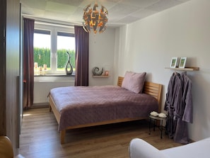 Ein Schlafzimmer mit Bett, Nachttisch, Stuhl, Fenster mit Vorhängen, einer Chandelierleuchtre und einem Regal mit Bildern.