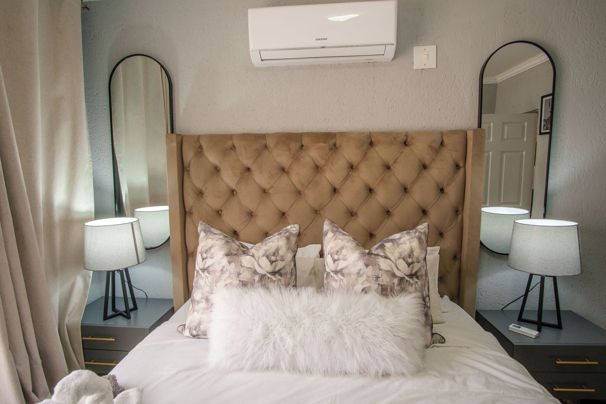 Un dormitorio con cabecera capitonada, almohadas florales, una manta blanca mullida, lámparas de noche y aire acondicionado de pared.