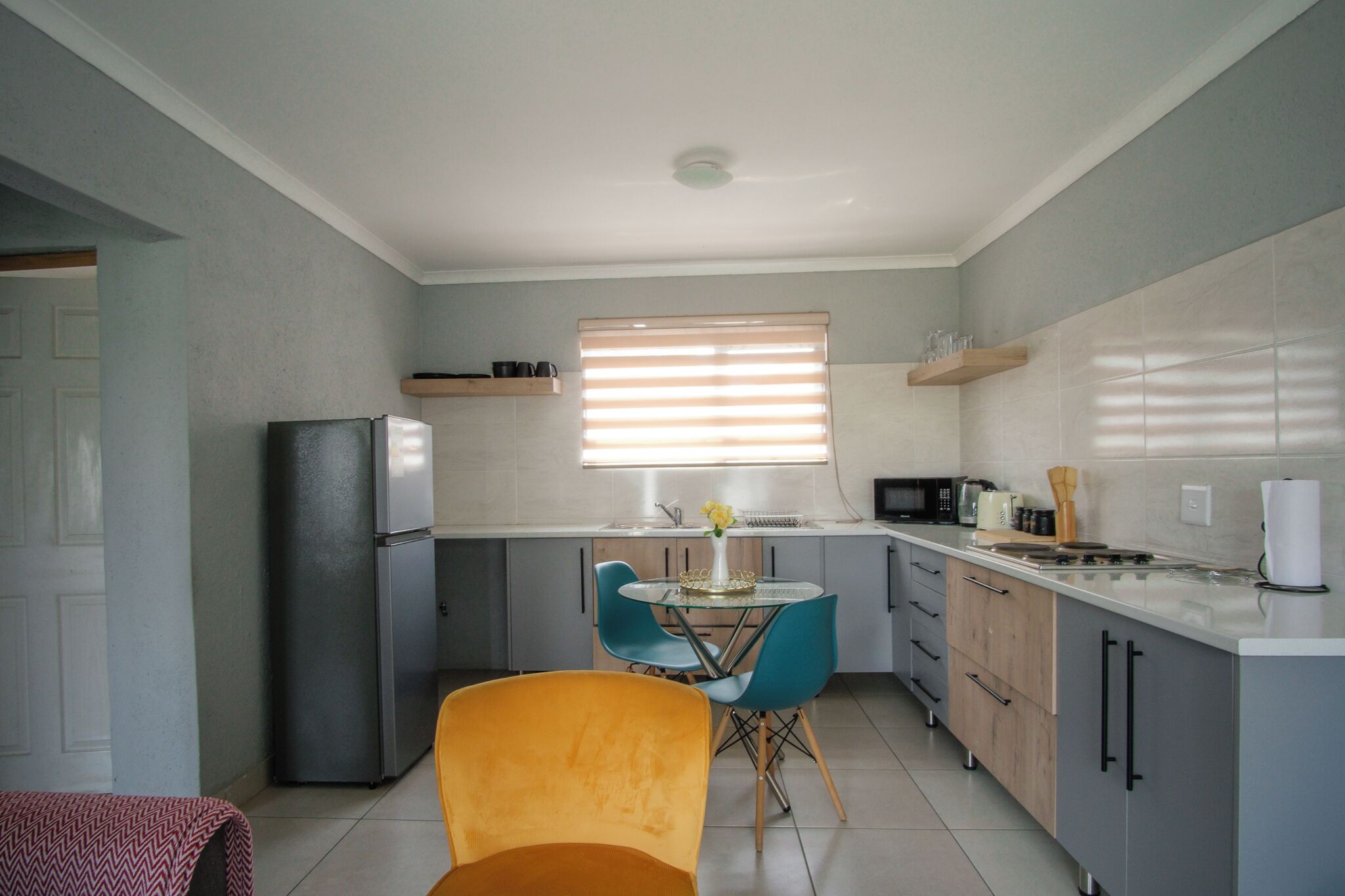 Een kleine keuken met een koelkast, magnetron en gootsteen; een eettafel met stoelen; en een raam met jaloezieën.