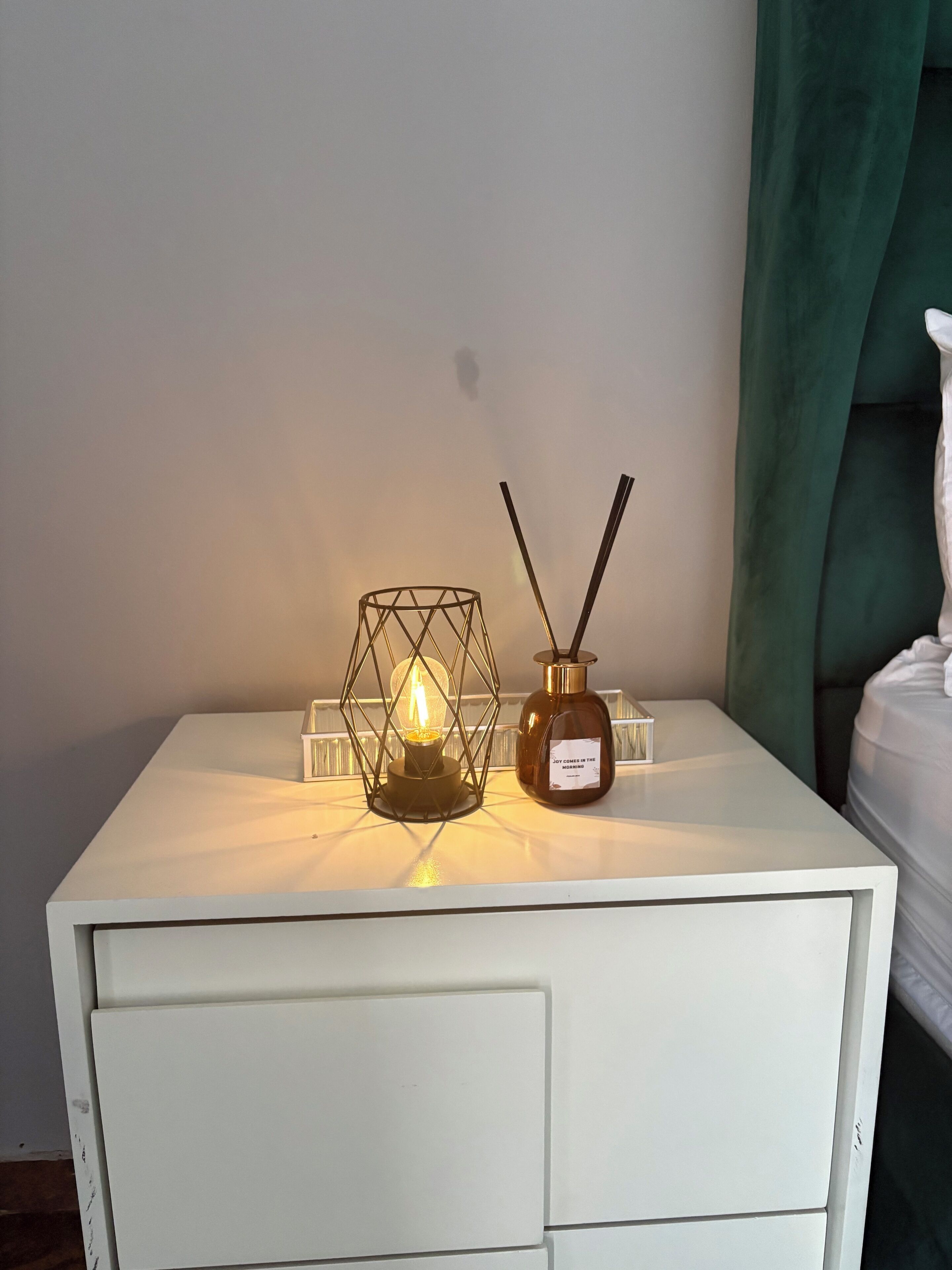 A white bedside table with a lit candle in a geometric holder, a bottle of essential oil, and a pair of reed diffusers.