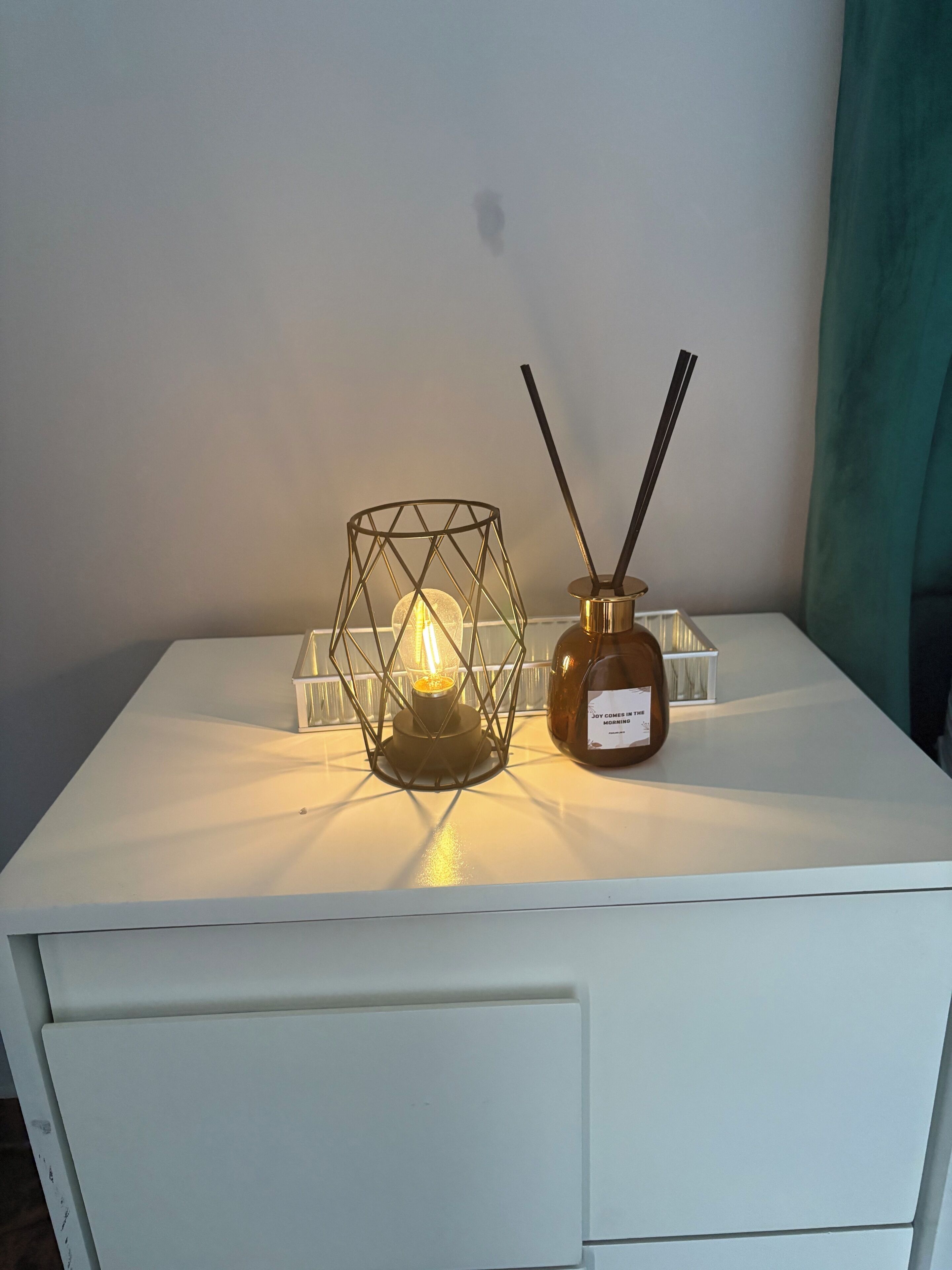 A white table with a geometric glass candle holder containing a lit candle, a small bottle with a label, and two black sticks.