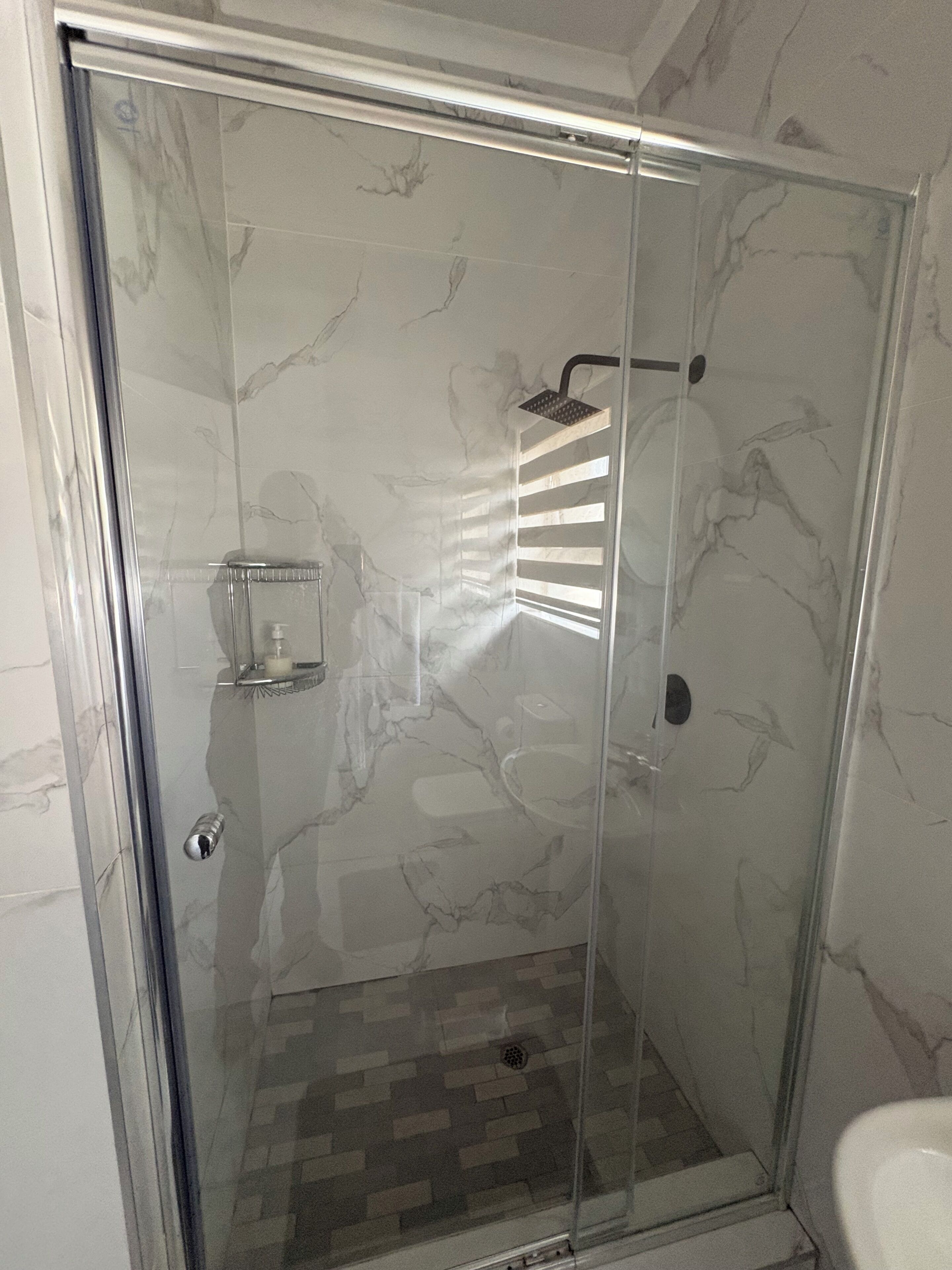 A glass-enclosed shower with a marble wall, a handheld showerhead, and a soap dispenser.